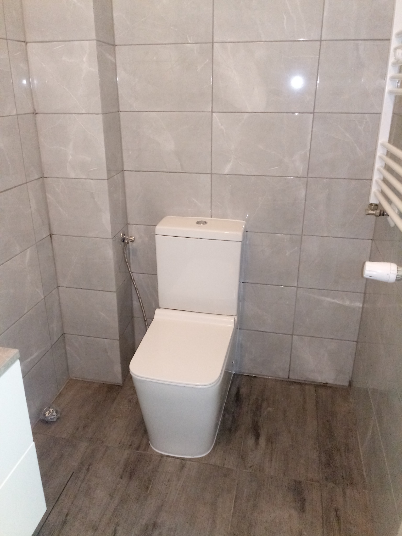 Nowoczesna toaleta w łazience wykończonej szarymi płytkami imitującymi marmur i panelami podłogowymi w kolorze drewna, widoczny grzejnik drabinkowy.