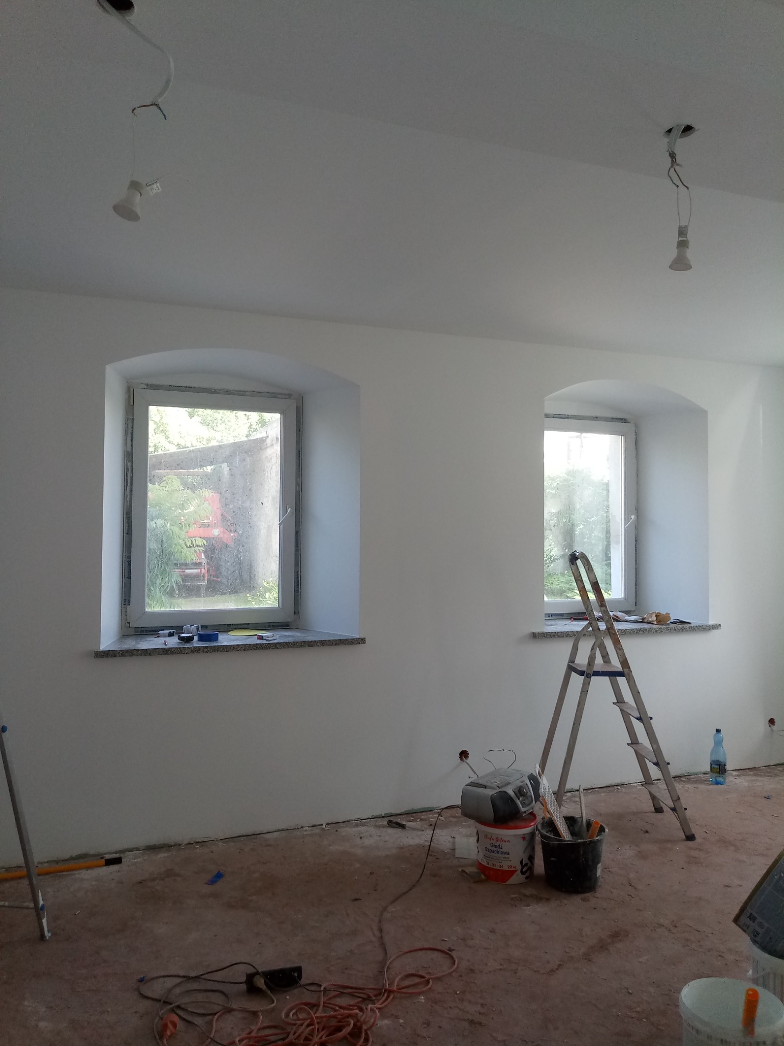 Wnętrze pokoju z dwoma oknami o łukowatym kształcie, ściany świeżo pomalowane na biało, widoczne ślady prac na podłodze, drabina oparta o ścianę, wiadro z narzędziami i puszki z farbą, brakujące...