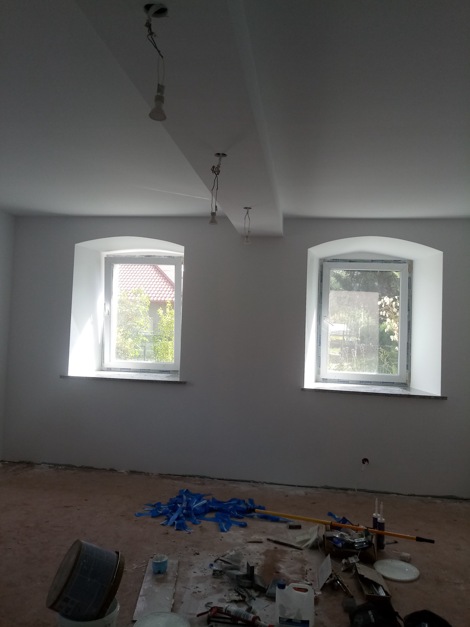 Wnętrze pokoju po świeżym malowaniu ścian na biało, dwa okna z zaokrąglonymi łukami, widoczne narzędzia i resztki materiałów malarskich na podłodze.