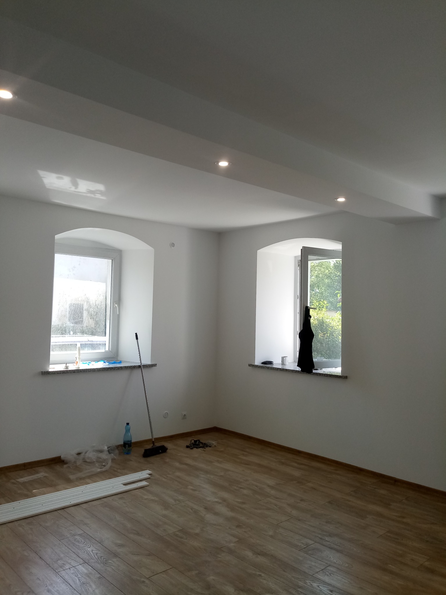 Wnętrze pokoju po remoncie z drewnianą podłogą, białymi ścianami i dwoma oknami o łukowatych framugach, widoczne listwy przypodłogowe, mop i butelka wody.