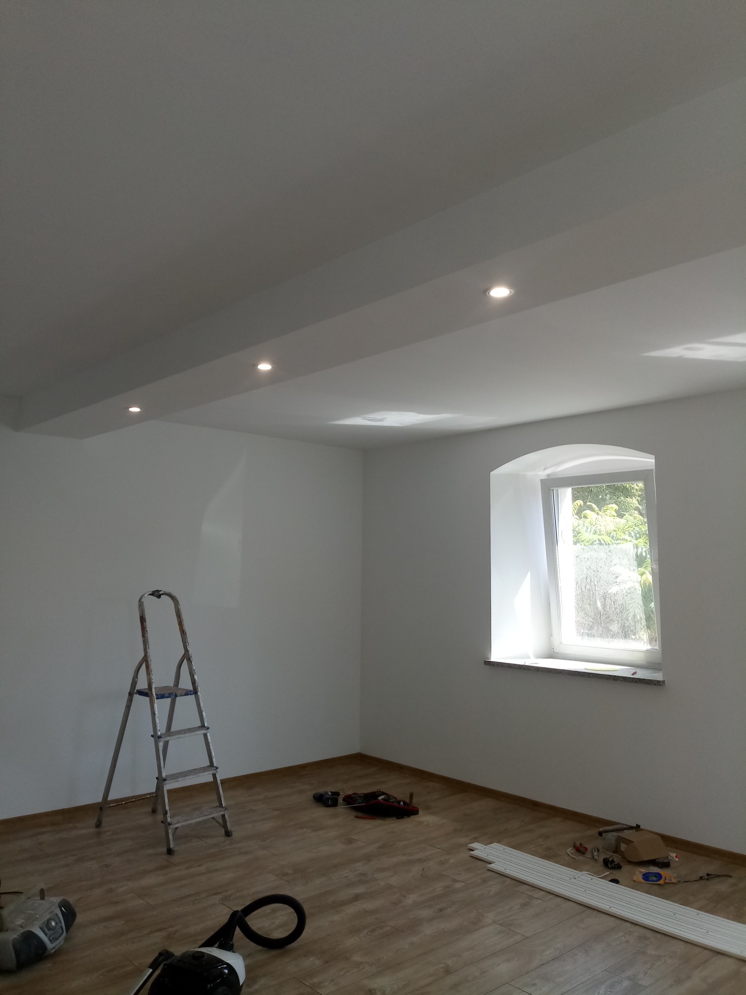 Pomalowane na biało wnętrze pokoju z oknem w kształcie łuku, drewnianą podłogą, aluminiową drabiną i elementami wykończeniowymi na podłodze, przygotowane do dalszych prac.
