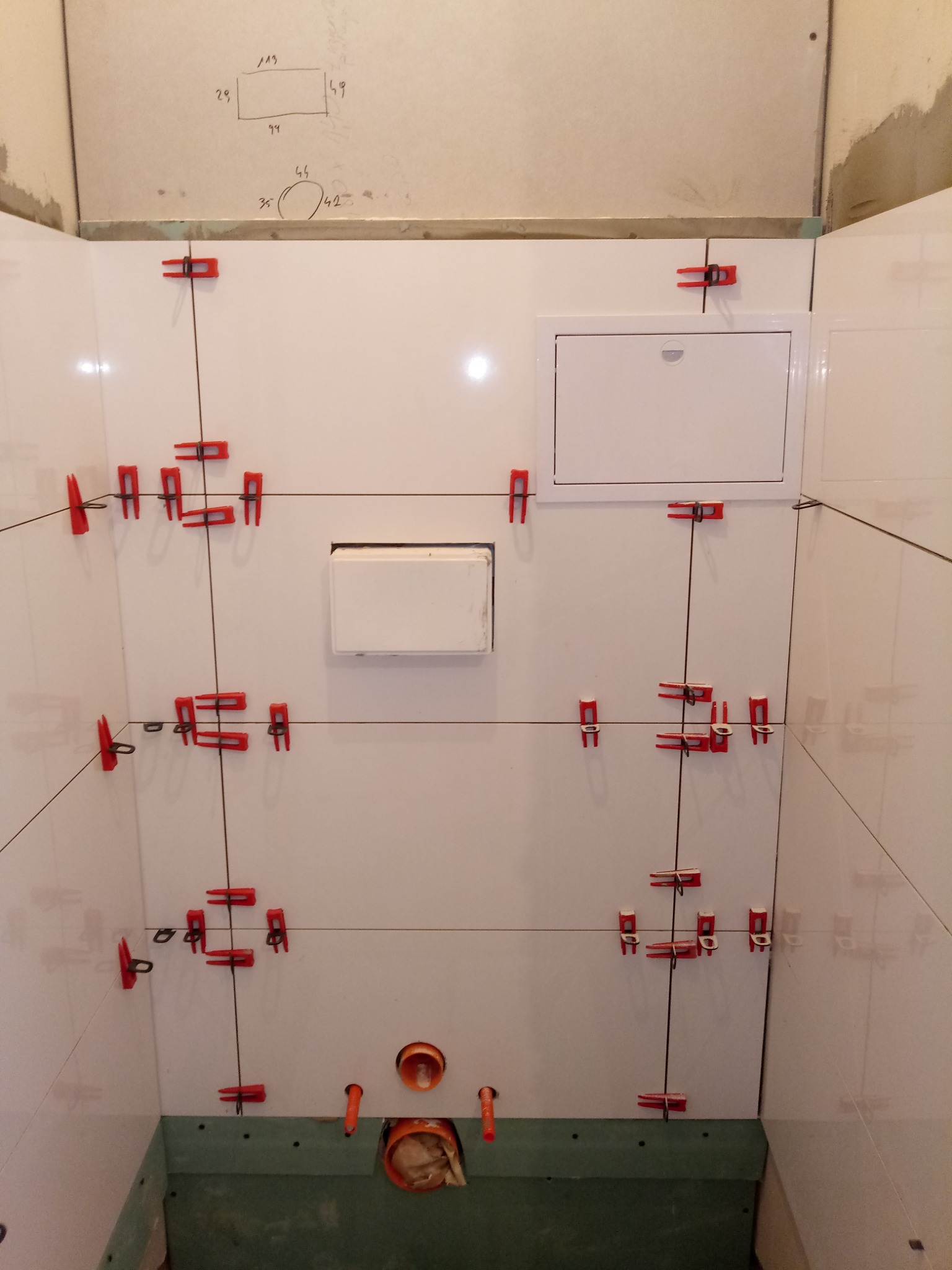 Białe płytki ścienne w łazience w trakcie montażu, z czerwonymi klipsami poziomującymi i widocznymi instalacjami hydraulicznymi u dołu.
