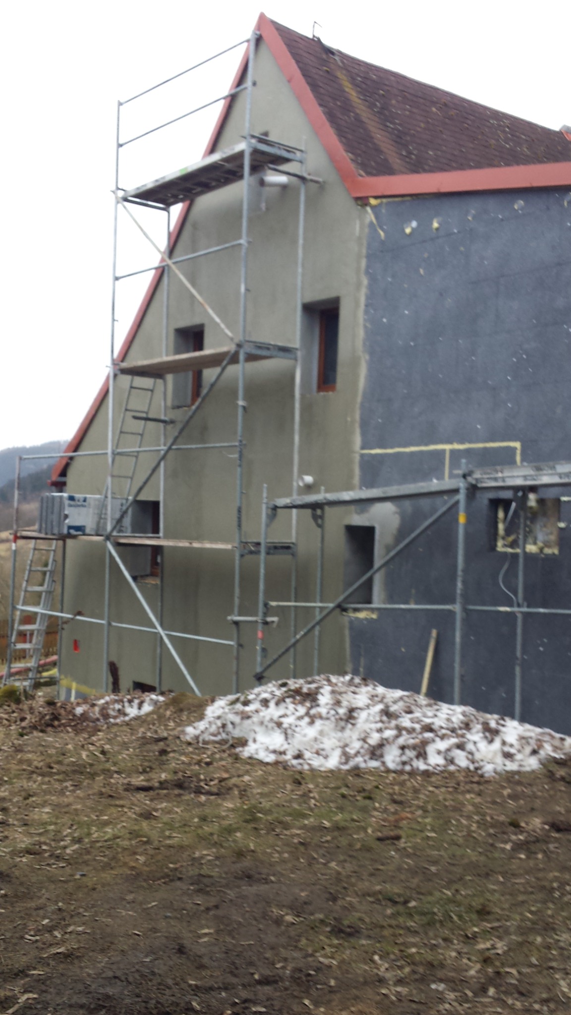 Dom w trakcie prac elewacyjnych: jedna ściana ocieplona i pokryta szarą płytą, druga w trakcie szpachlowania, rusztowania ustawione przy ścianach, w tle las.