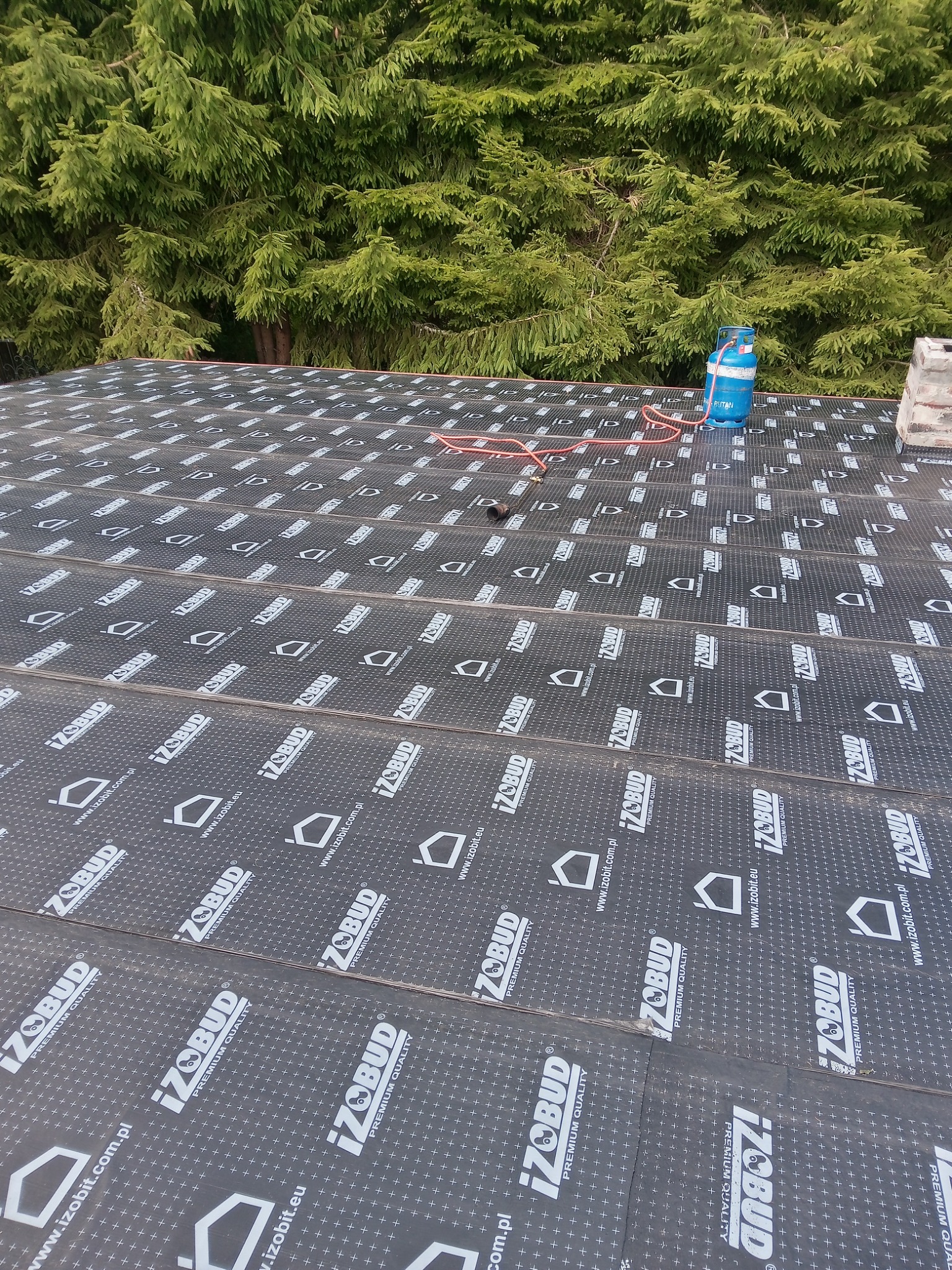 Widok z góry na fragment dachu pokrytego czarną papą z nadrukiem IzoBud, w tle zielone drzewa iglaste i niebieska butla gazowa z czerwoną rurką.