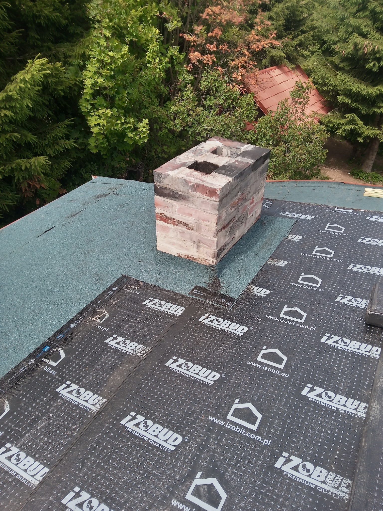 Widok z góry na fragment dachu pokrytego papą z wyraźnym kominem z cegły i zielonymi drzewami w tle.