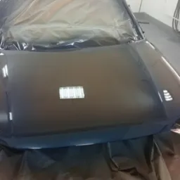 Błyszcząca, świeżo polakierowana maska samochodu w kolorze ciemnego granatu, zabezpieczona folią ochronną w lakierni, z widocznym odbiciem lamp sufitowych i drewnianym stołkiem w tle.