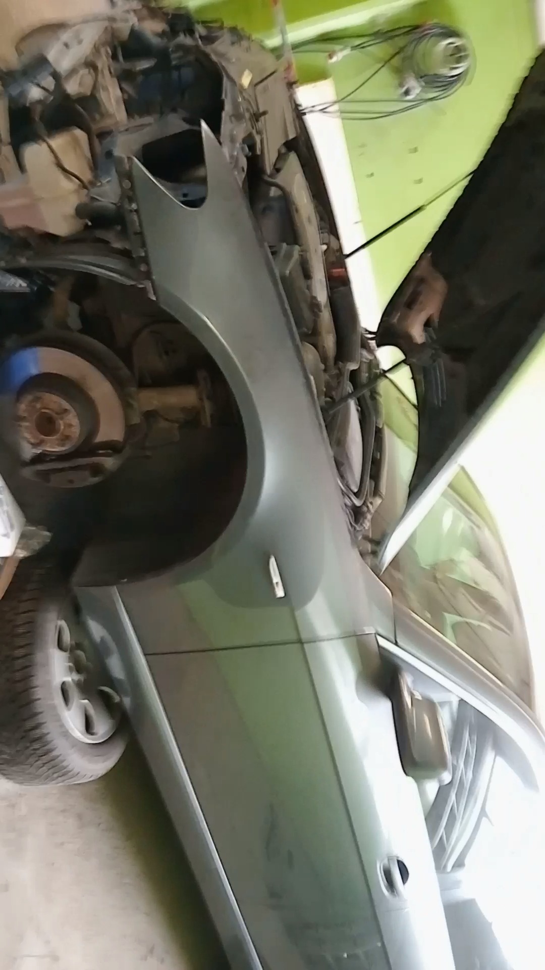 Szary samochód z otwartą maską, widoczny fragment koła, błotnik i wnętrze komory silnika w warsztacie.