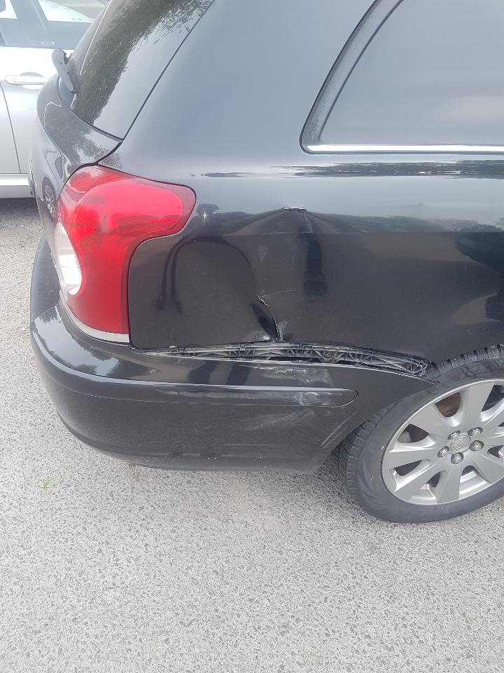 Uszkodzony tył czarnego samochodu typu kombi. Widoczne wgniecenie na błotniku oraz pęknięty plastikowy element pod lampą. Opona lekko zabrudzona.