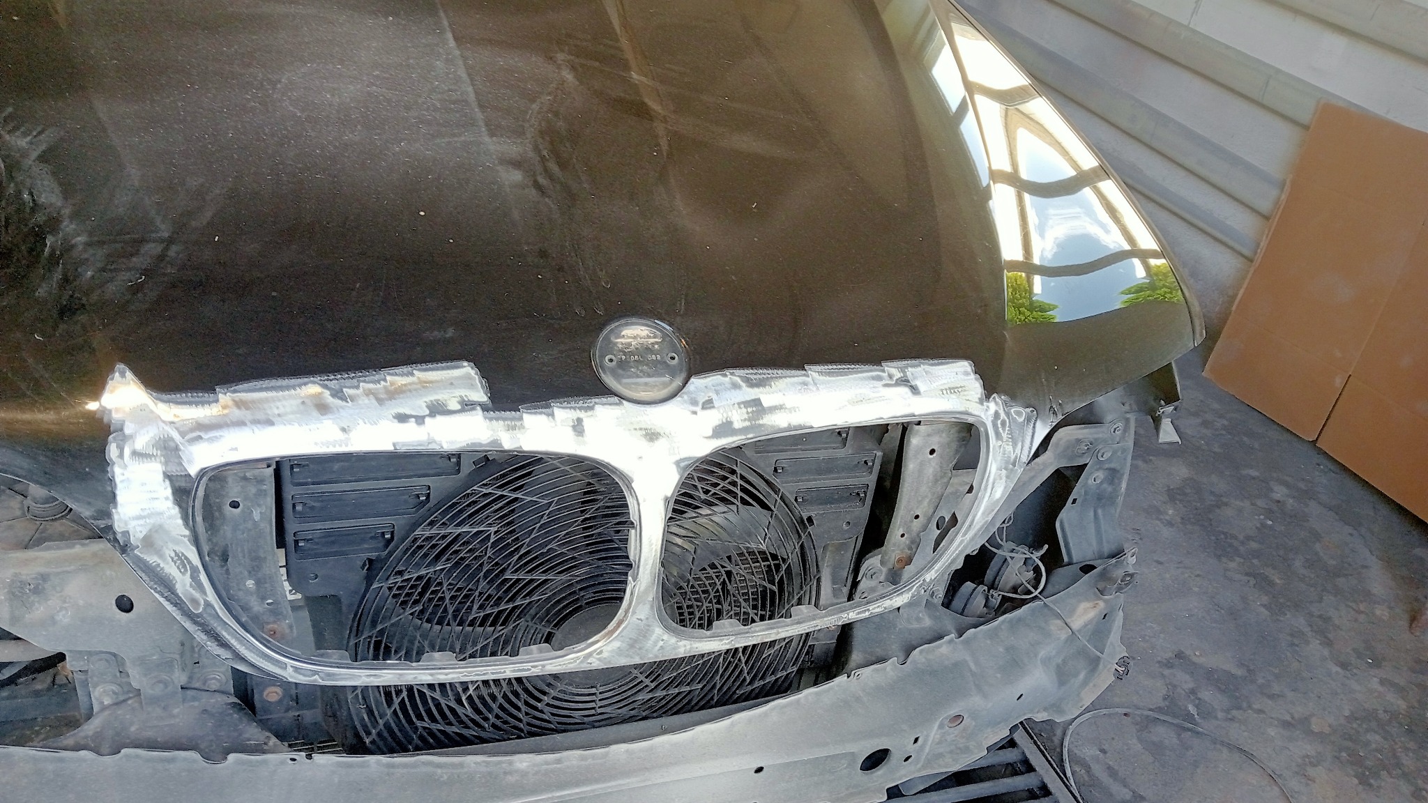 Czarny samochód BMW z uszkodzonym przodem, w trakcie naprawy blacharskiej, widoczne ślady szpachli i spawania wokół atrapy chłodnicy.