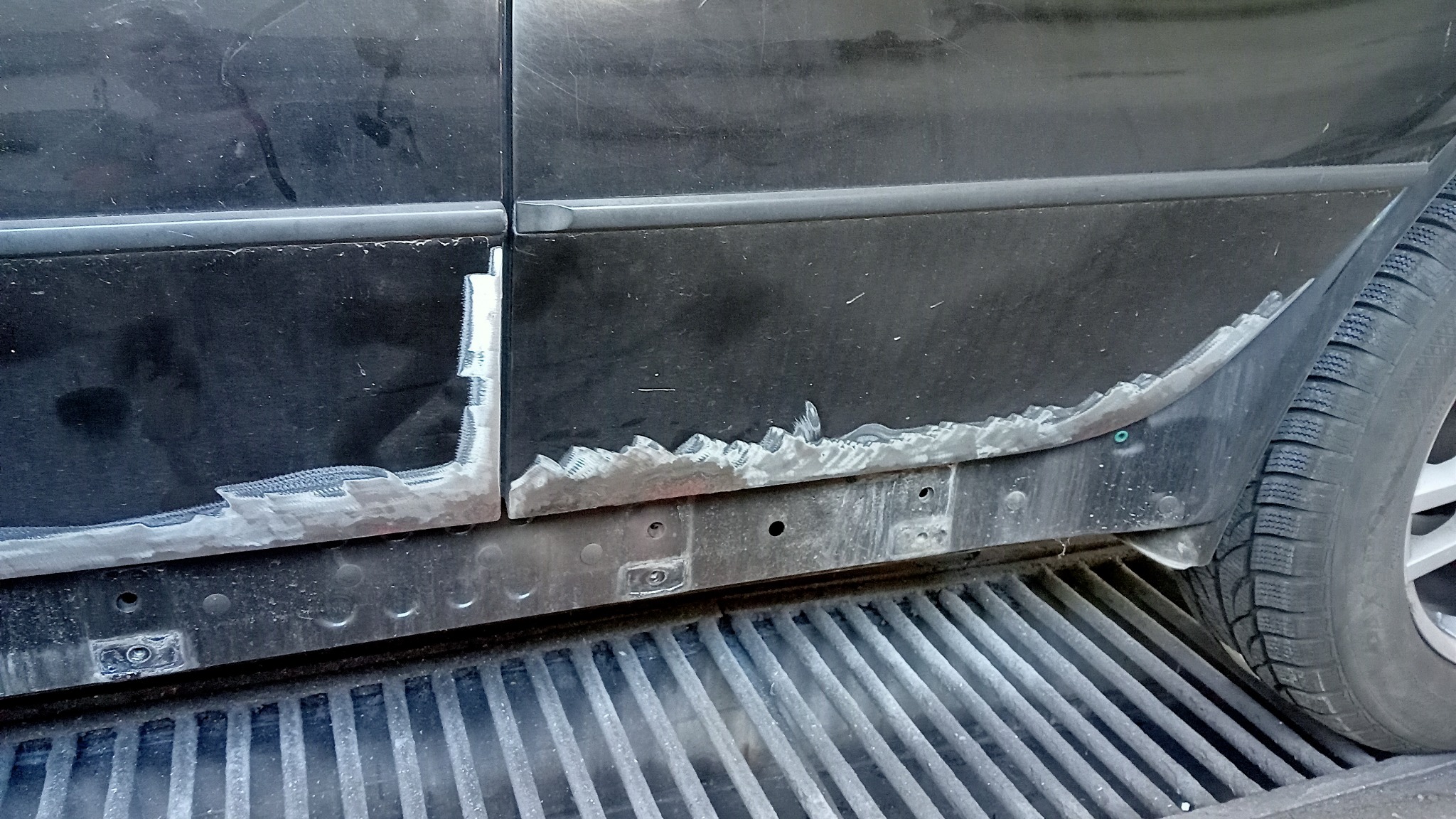 Fragment karoserii czarnego samochodu z widocznymi śladami napraw blacharskich, w tym obszar zgrzewania i spawania, na tle kratki ściekowej.