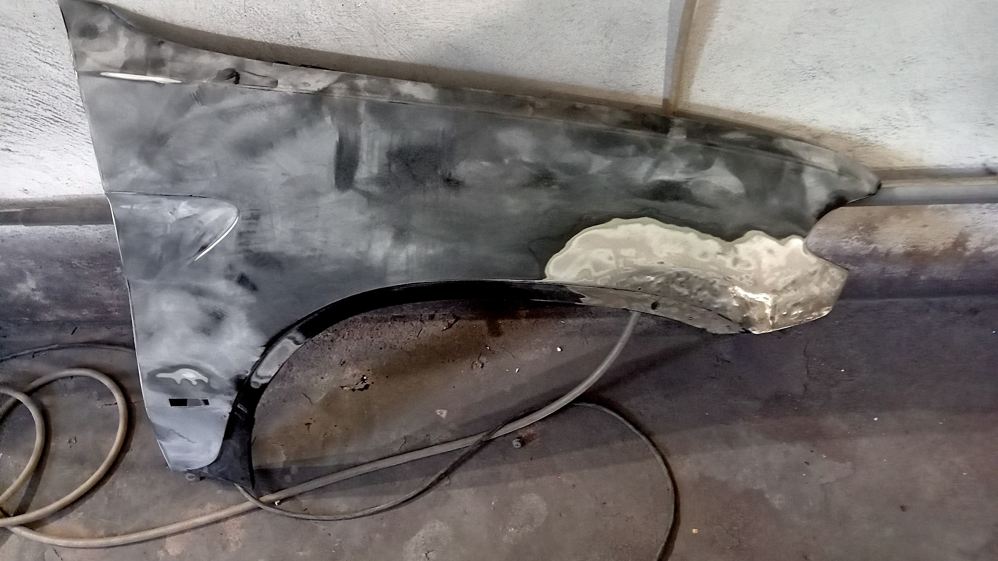 Błotnik samochodowy w trakcie naprawy blacharskiej, z widocznymi śladami szpachli i przygotowania do lakierowania, leżący na podłodze warsztatu.