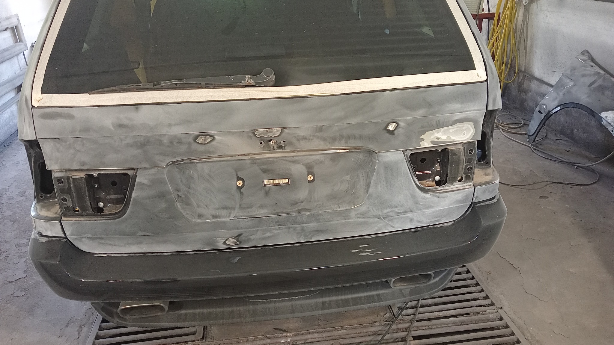 Tył samochodu kombi przygotowany do lakierowania, z widocznymi szpachlowanymi miejscami, taśmą maskującą wokół szyby i brakującymi lampami, w warsztacie blacharskim.