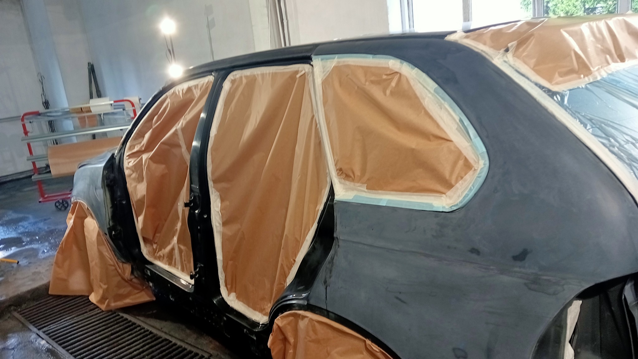 Samochód osobowy przygotowany do lakierowania, z zaklejonymi oknami i zabezpieczonymi elementami karoserii papierem maskującym oraz taśmą malarską, widoczne ślady szpachlowania.