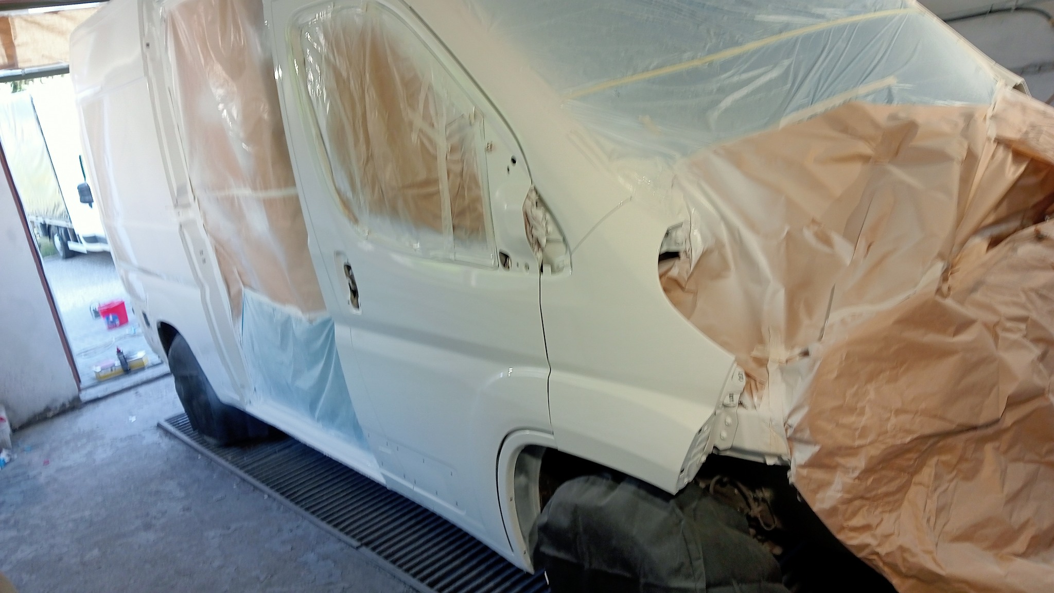 Biały samochód dostawczy w trakcie lakierowania, zabezpieczony folią i papierem maskującym, widoczny na kratce wentylacyjnej w warsztacie.