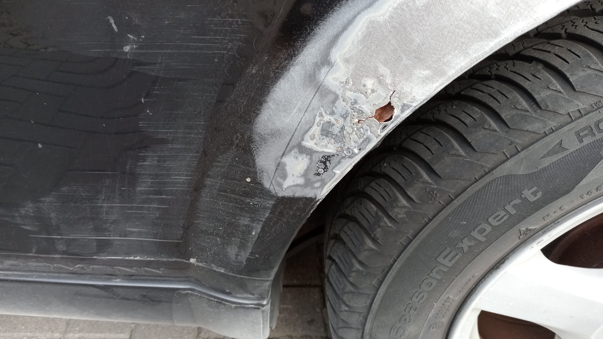 Fragment czarnego samochodu z widoczną korozją na błotniku, przygotowanego do naprawy blacharskiej, obok opona zimowa.