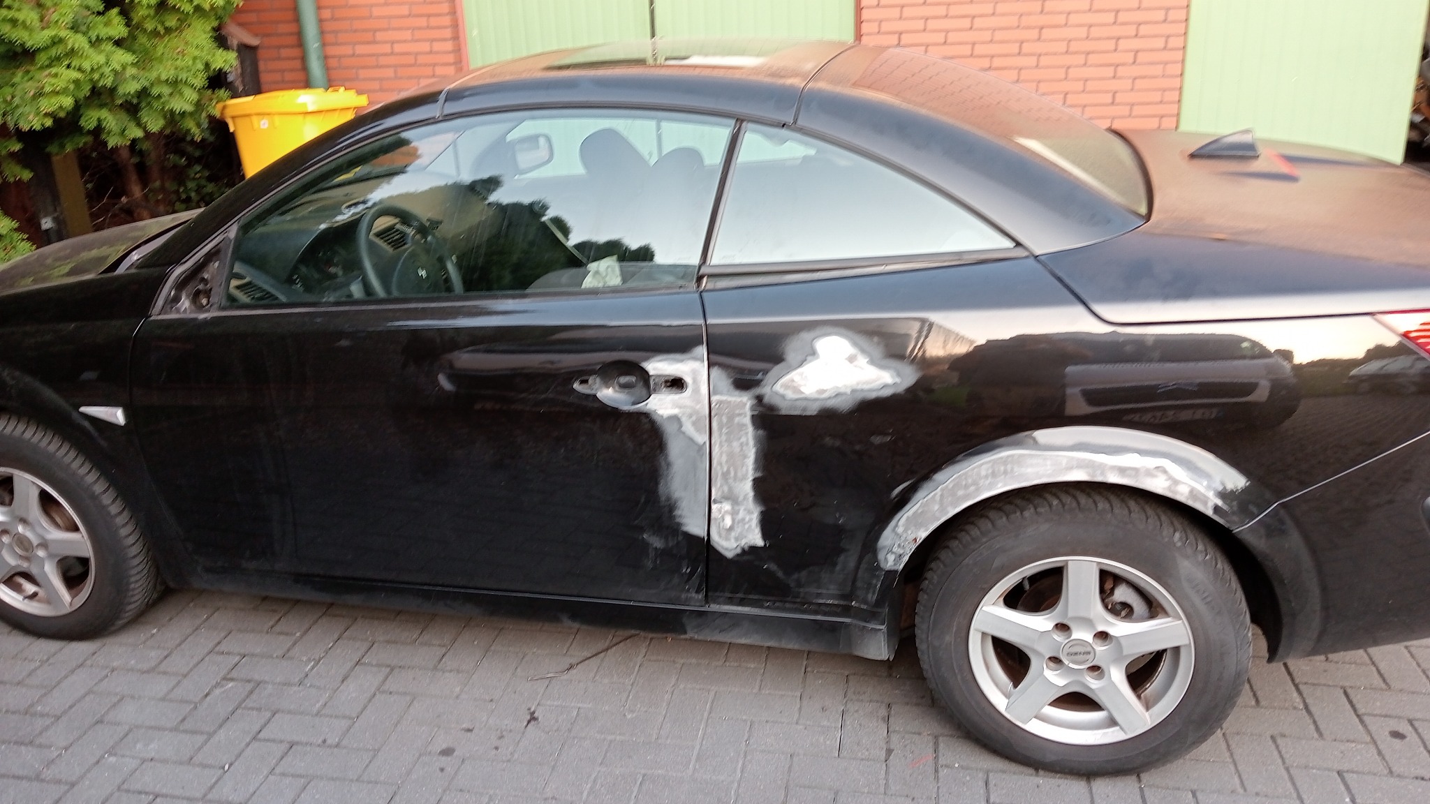 Czarny samochód typu kabriolet z widocznymi śladami napraw blacharskich, szpachlą na drzwiach i nadkolu, stojący na kostce brukowej przed budynkiem z cegły.