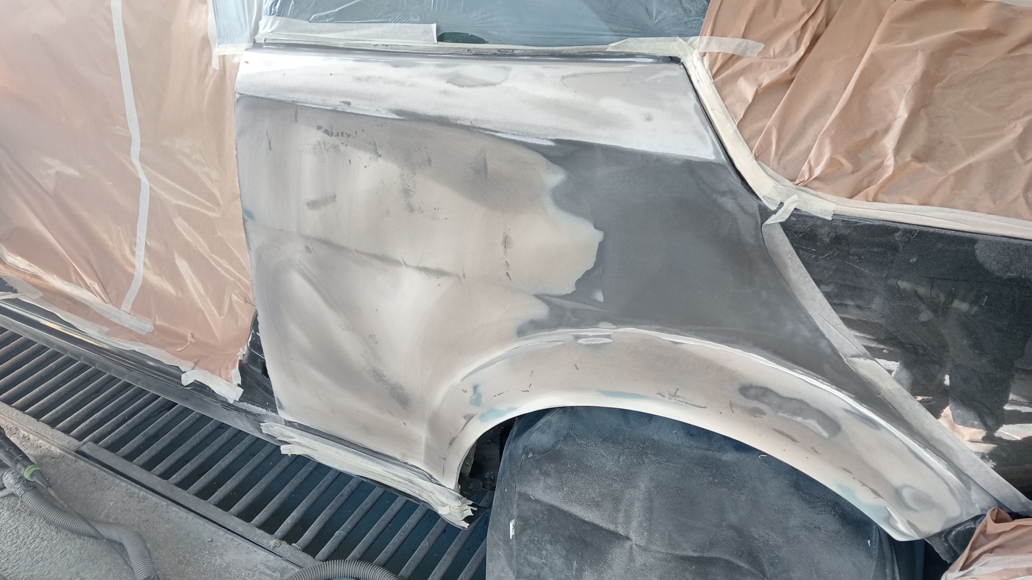 Przygotowanie elementu karoserii samochodu do lakierowania: szpachlowany błotnik, zabezpieczony folią i taśmą, widoczna kratka odpływowa w warsztacie.