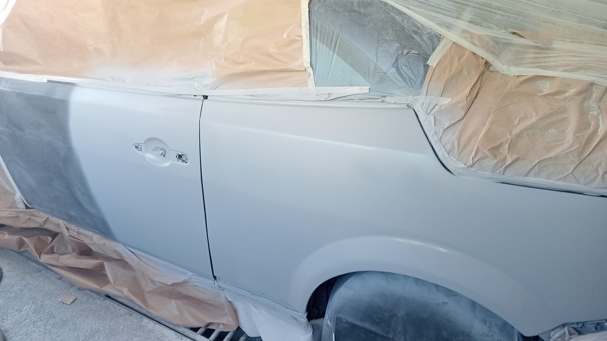 Biała karoseria samochodu częściowo zakryta papierem maskującym i folią ochronną, przygotowana do lakierowania, widoczny fragment drzwi z klamką i nadkole.