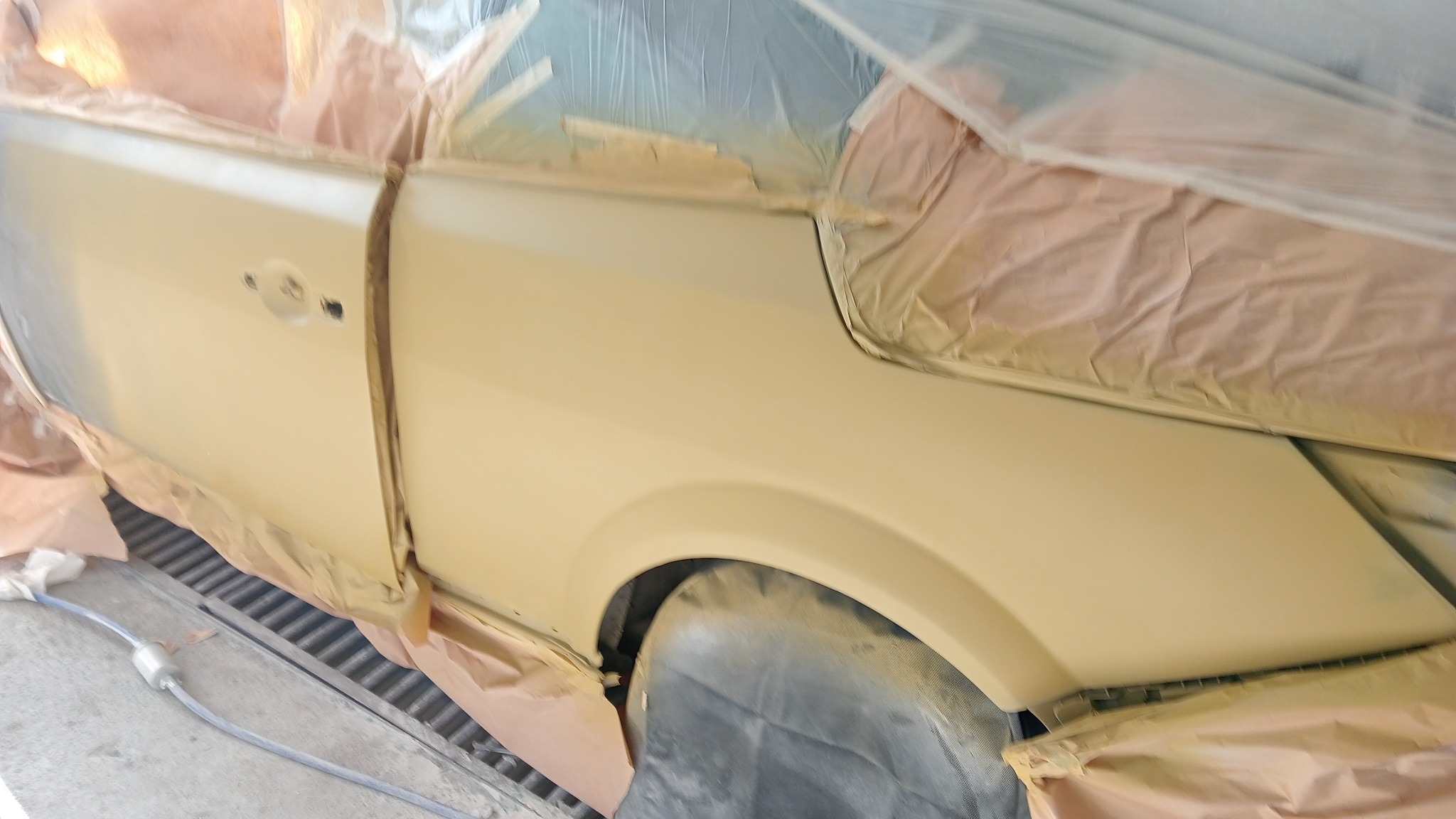 Samochód w trakcie lakierowania, zabezpieczony folią i papierem, świeżo pokryty podkładem w kolorze beżowym.