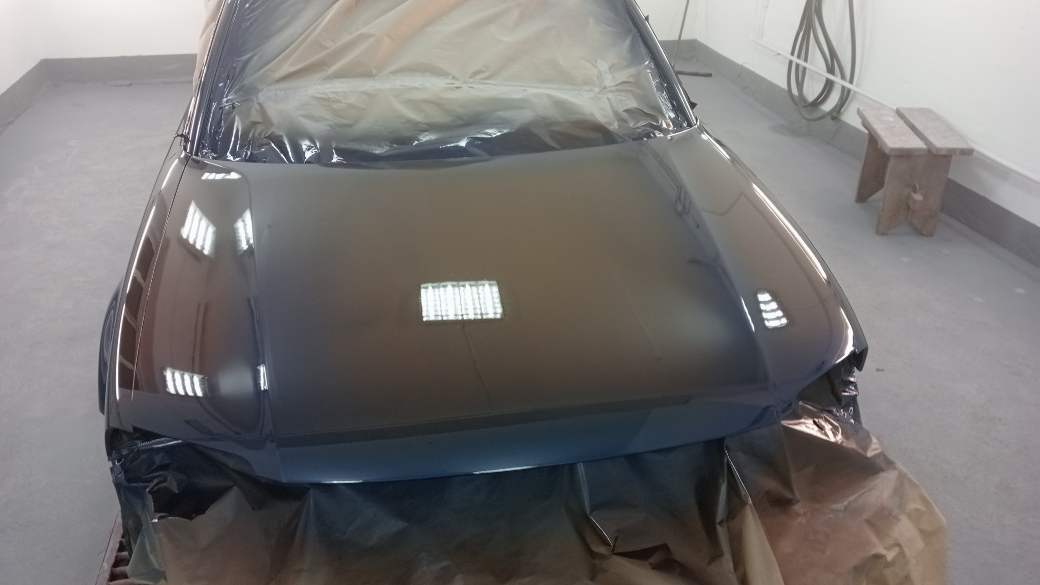 Błyszcząca, świeżo polakierowana maska samochodu w kolorze ciemnego granatu, zabezpieczona folią ochronną w lakierni, z widocznym odbiciem lamp sufitowych i drewnianym stołkiem w tle.
