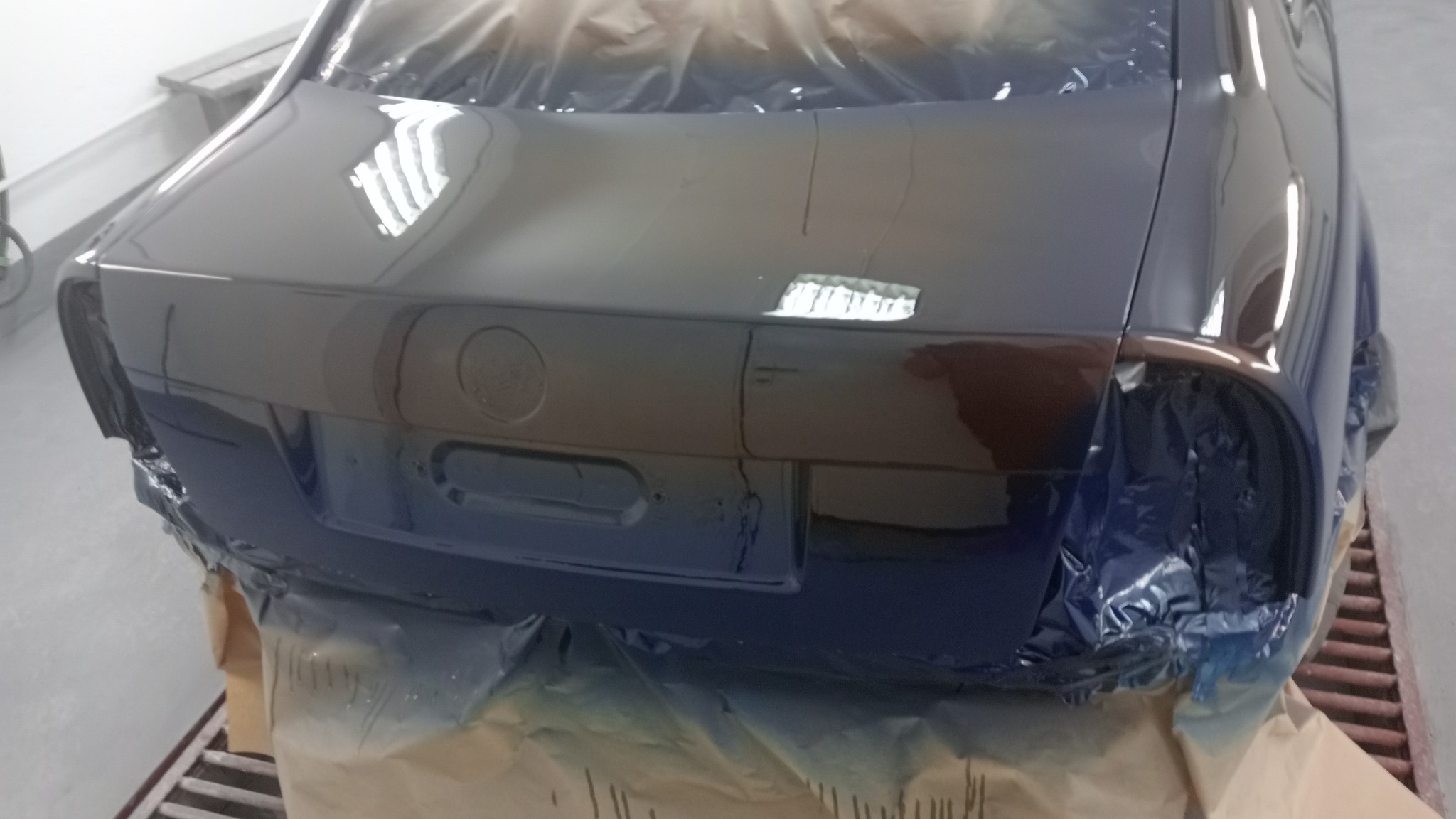 Tylna część samochodu w trakcie lakierowania na kolor granatowy, zabezpieczona folią malarską, z widocznymi refleksami światła na świeżej powłoce lakierniczej.