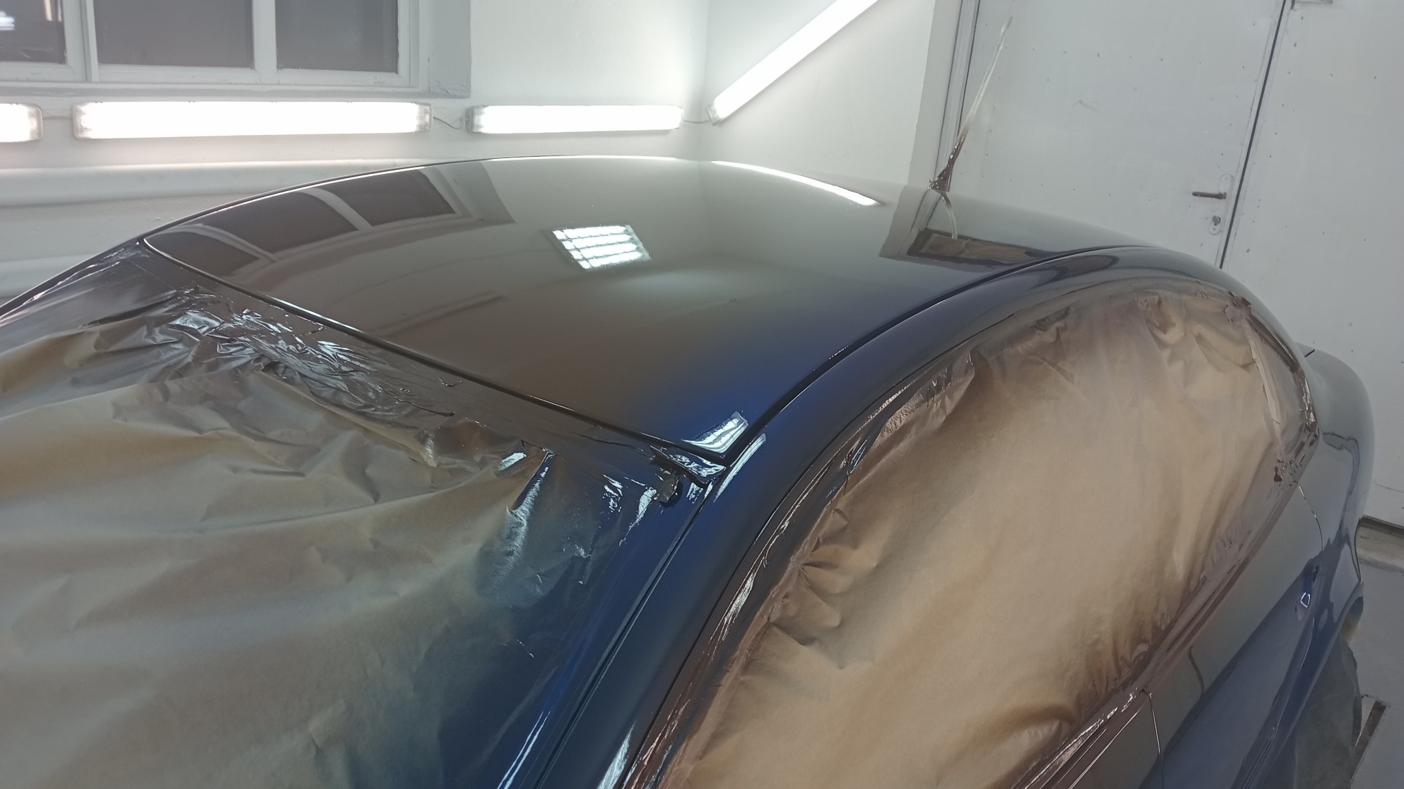 Błyszczący, granatowy dach samochodu po lakierowaniu, częściowo zakryty papierem maskującym w jasnym warsztacie.