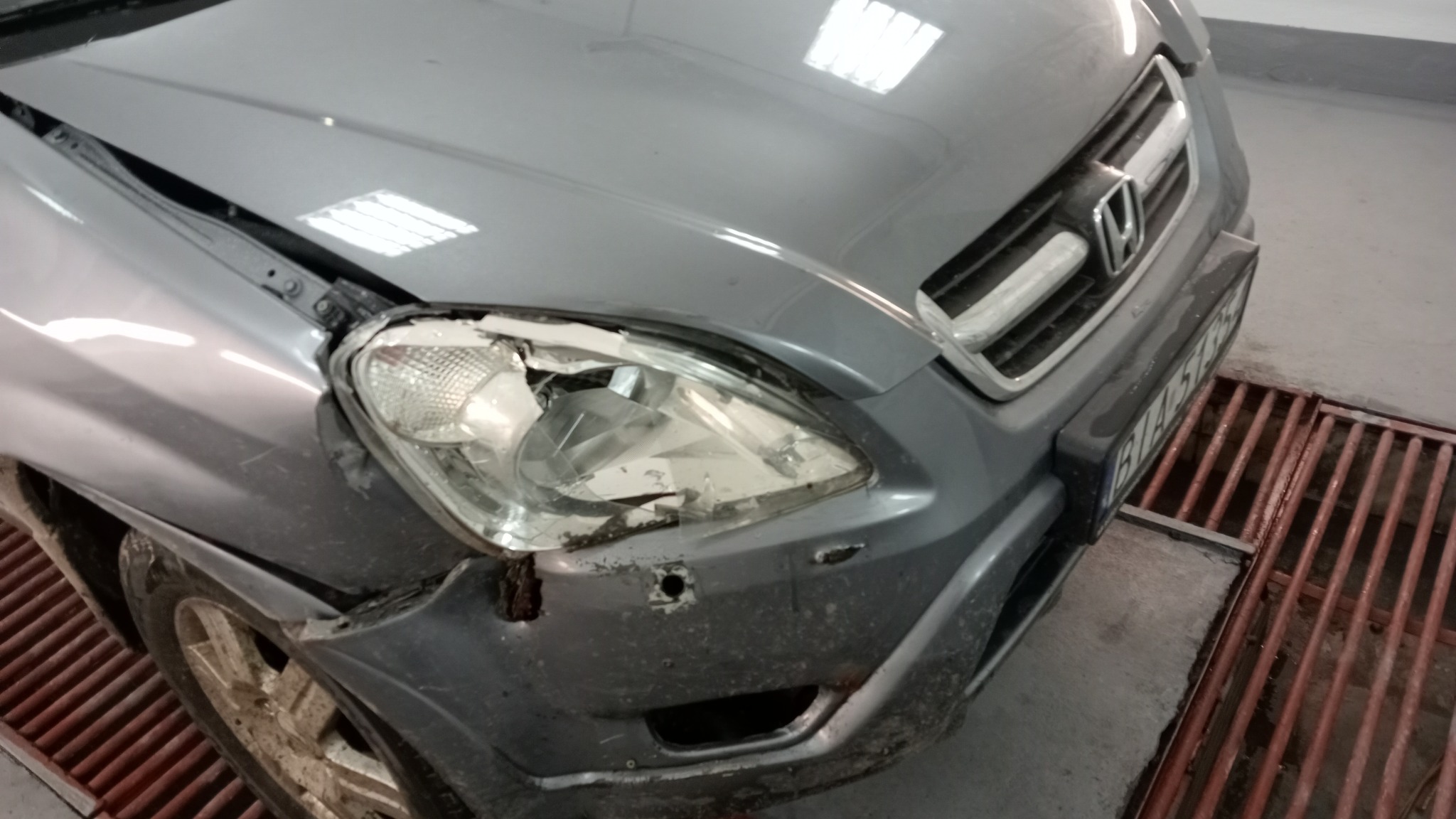 Uszkodzony przód srebrnego samochodu Honda CR-V z rozbitym reflektorem i wgniecionym błotnikiem, widoczne ślady korozji, na podłożu kratka ściekowa.
