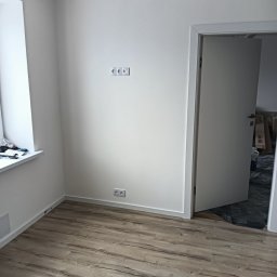 EFEKT KRZYSZTOF JANUSZ - Wnętrze pokoju po odświeżeniu, z jasnymi ścianami, drewnopodobną podłogą i otwartymi białymi drzwiami; widoczne gniazdka elektryczne i narzędzia malarskie na parapecie.