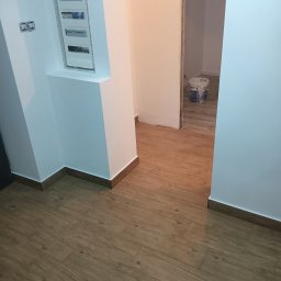 EFEKT KRZYSZTOF JANUSZ - Wnętrze po remoncie: świeżo pomalowane ściany na biało i niebiesko, drewnopodobne panele na podłodze, widoczna puszka z farbą w tle.