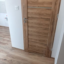 EFEKT KRZYSZTOF JANUSZ - Zamknięte, drewnopodobne drzwi wewnętrzne z poziomymi panelami i srebrną klamką, dopasowane do paneli podłogowych w odcieniach brązu, widoczne na tle białej ściany z włącznikiem.