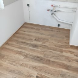 EFEKT KRZYSZTOF JANUSZ - Ułożone płytki podłogowe imitujące drewno w małej łazience z widocznymi rurami i instalacją elektryczną na ścianie.