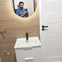 EFEKT KRZYSZTOF JANUSZ - Wykończona łazienka z umywalką na szafce i lustrem z podświetleniem LED, odbijającym osobę robiącą zdjęcie. Widoczne drewnopodobne płytki i białe drzwi.