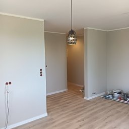 EFEKT KRZYSZTOF JANUSZ - Jasne, odświeżone wnętrze po malowaniu ścian na szaro, z drewnianą podłogą i nowoczesną lampą wiszącą. Widoczne puszki elektryczne bez osprzętu i narzędzia malarskie.