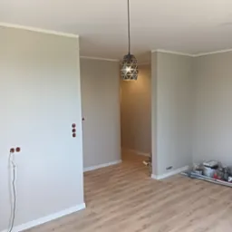 Jasne, odświeżone wnętrze po malowaniu ścian na szaro, z drewnianą podłogą i nowoczesną lampą wiszącą. Widoczne puszki elektryczne bez osprzętu i narzędzia malarskie.