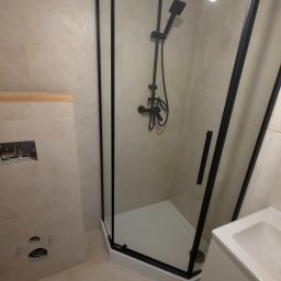 EFEKT KRZYSZTOF JANUSZ - Nowoczesna kabina prysznicowa z czarnymi profilami i deszczownicą, jasne płytki, fragment umywalki. Widoczny etap prac wykończeniowych w łazience.