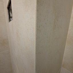 EFEKT KRZYSZTOF JANUSZ - Ściana łazienki wyłożona beżowymi płytkami z widocznym otworem na instalację. Widoczny narożnik i fugi. Delikatna faktura płytek. Ujęcie z dołu.