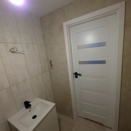 EFEKT KRZYSZTOF JANUSZ - Wykończona łazienka z beżowymi płytkami, białą szafką z umywalką i czarną armaturą. Widoczne białe drzwi z przeszkleniami i wystające przewody.