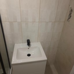 EFEKT KRZYSZTOF JANUSZ - Biała umywalka z czarną baterią w wykończonej łazience z beżowymi, pionowymi płytkami. Widoczne fragmenty kabiny prysznicowej i niedokończone gniazdko elektryczne.