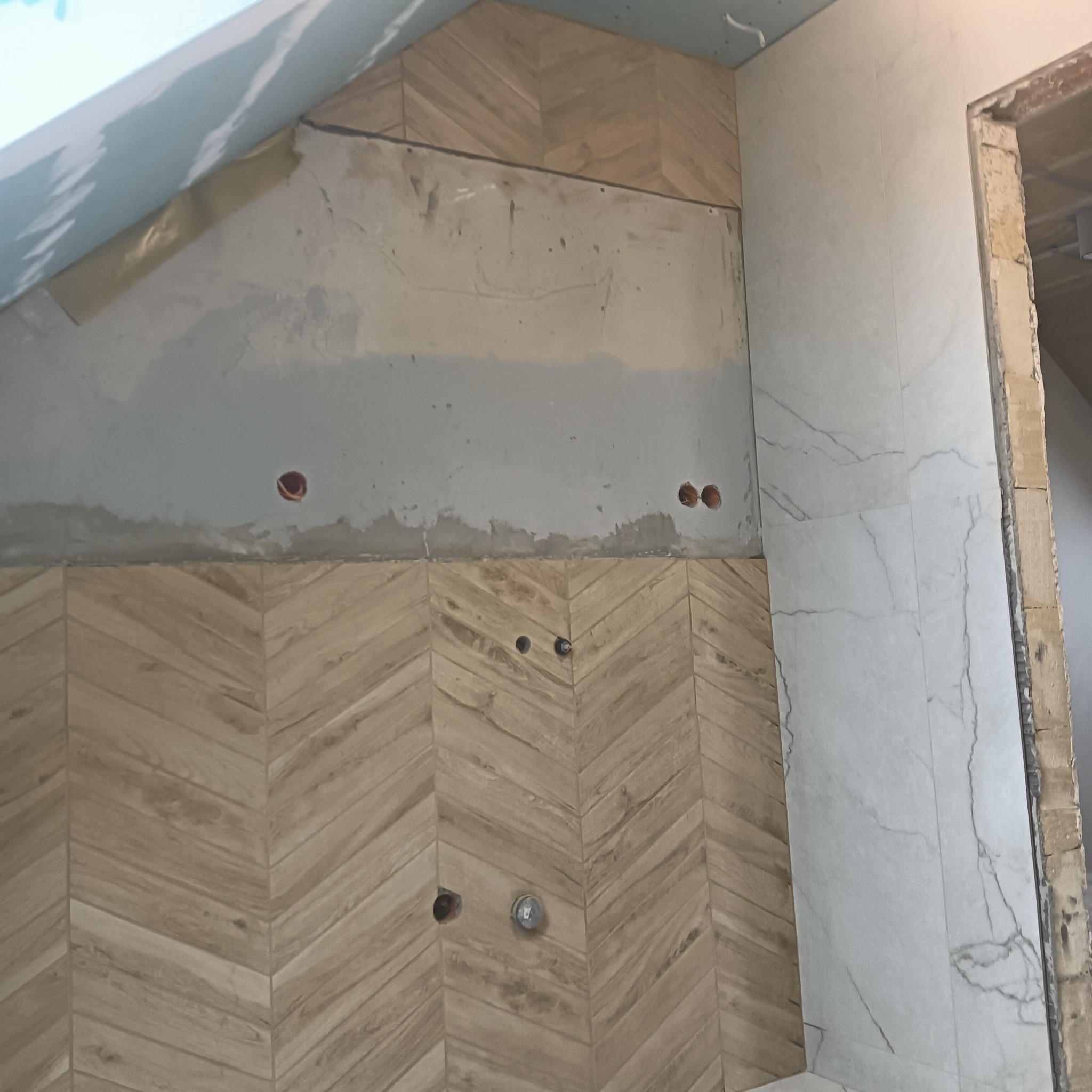 Montaż płytek drewnopodobnych w jodełkę na ścianie łazienki, widoczne otwory na instalacje oraz fragment ściany przygotowanej pod dalsze prace wykończeniowe.
