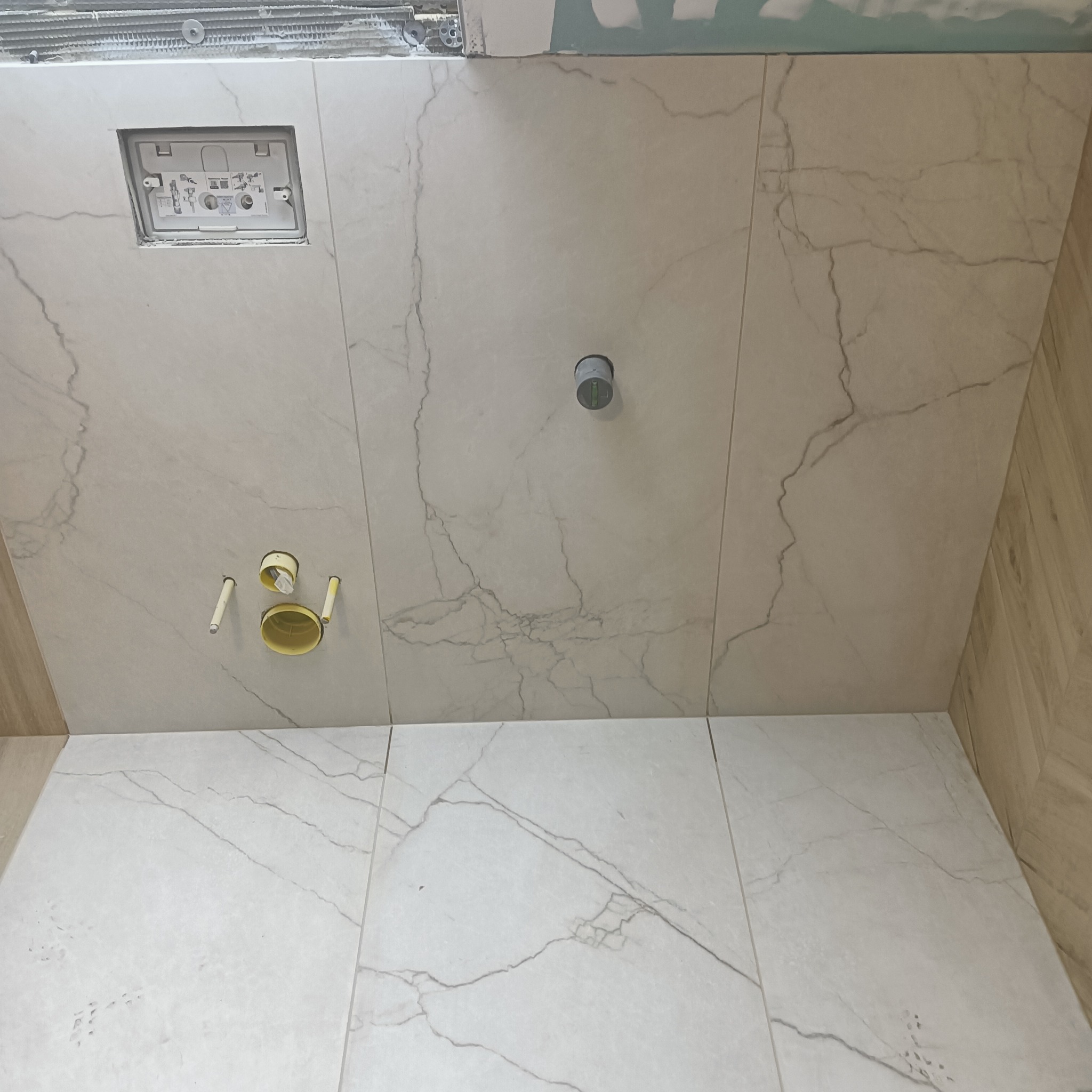 Ściana i podłoga w łazience wyłożone płytkami imitującymi marmur, z widocznymi otworami na instalacje sanitarne oraz zamontowaną podtynkową spłuczką WC w trakcie instalacji.
