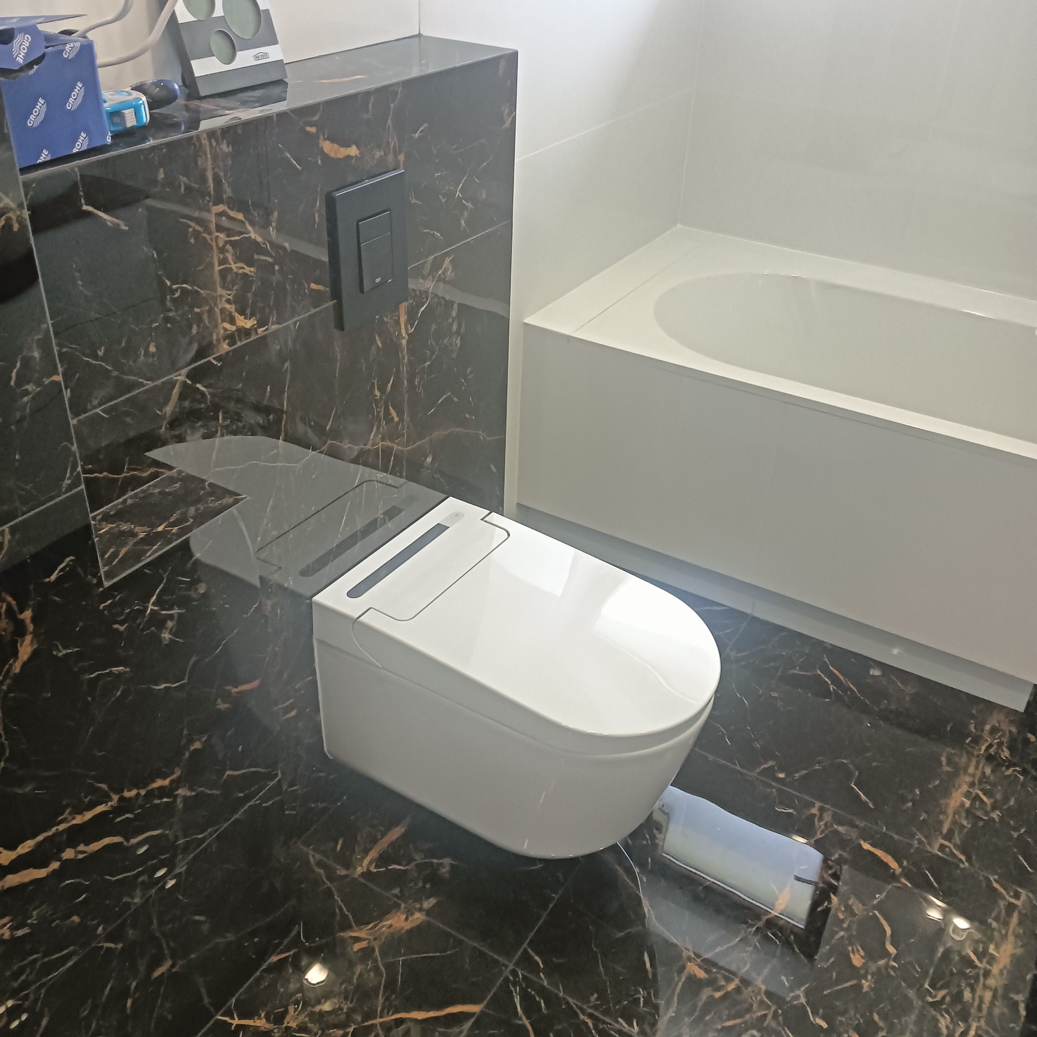 Nowoczesna toaleta myjąca z czarną, marmurkową zabudową i białą wanną w tle, widoczne pudełka i akcesoria instalacyjne na blacie.