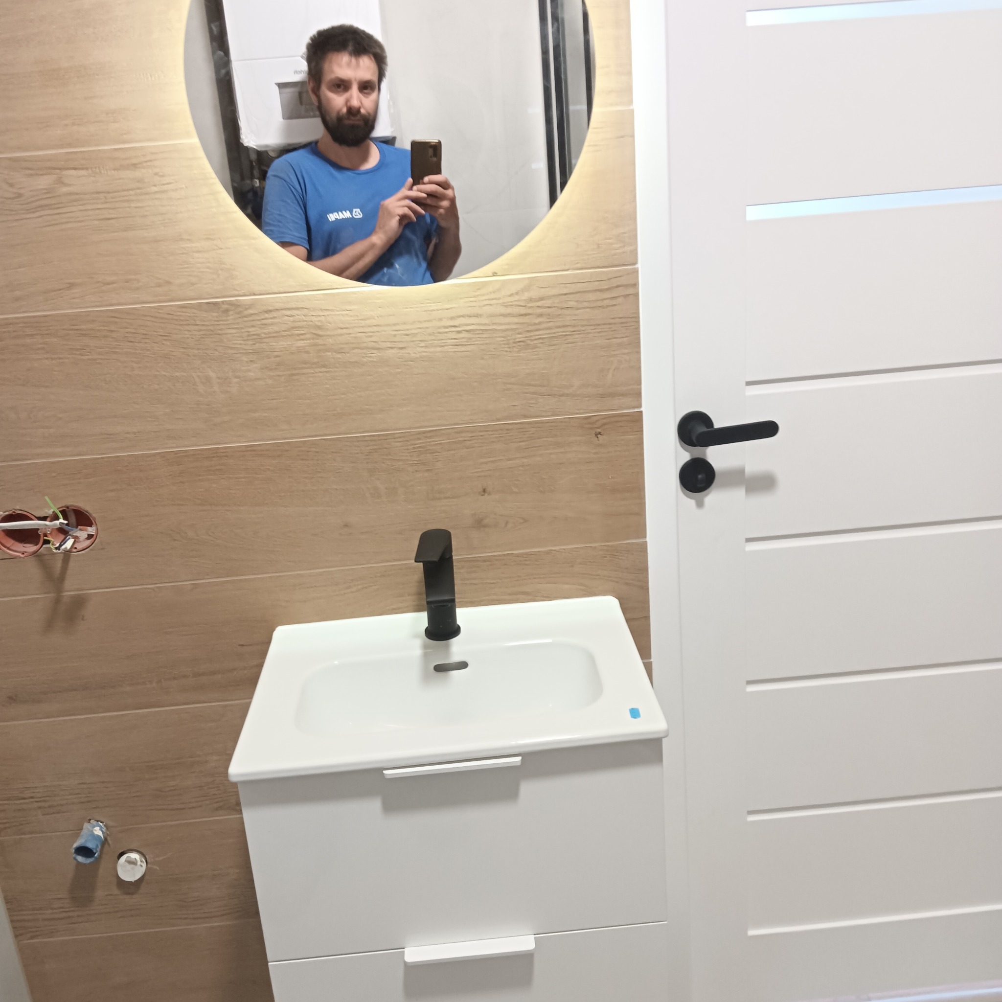 Wykończona łazienka z umywalką na szafce i lustrem z podświetleniem LED, odbijającym osobę robiącą zdjęcie. Widoczne drewnopodobne płytki i białe drzwi.