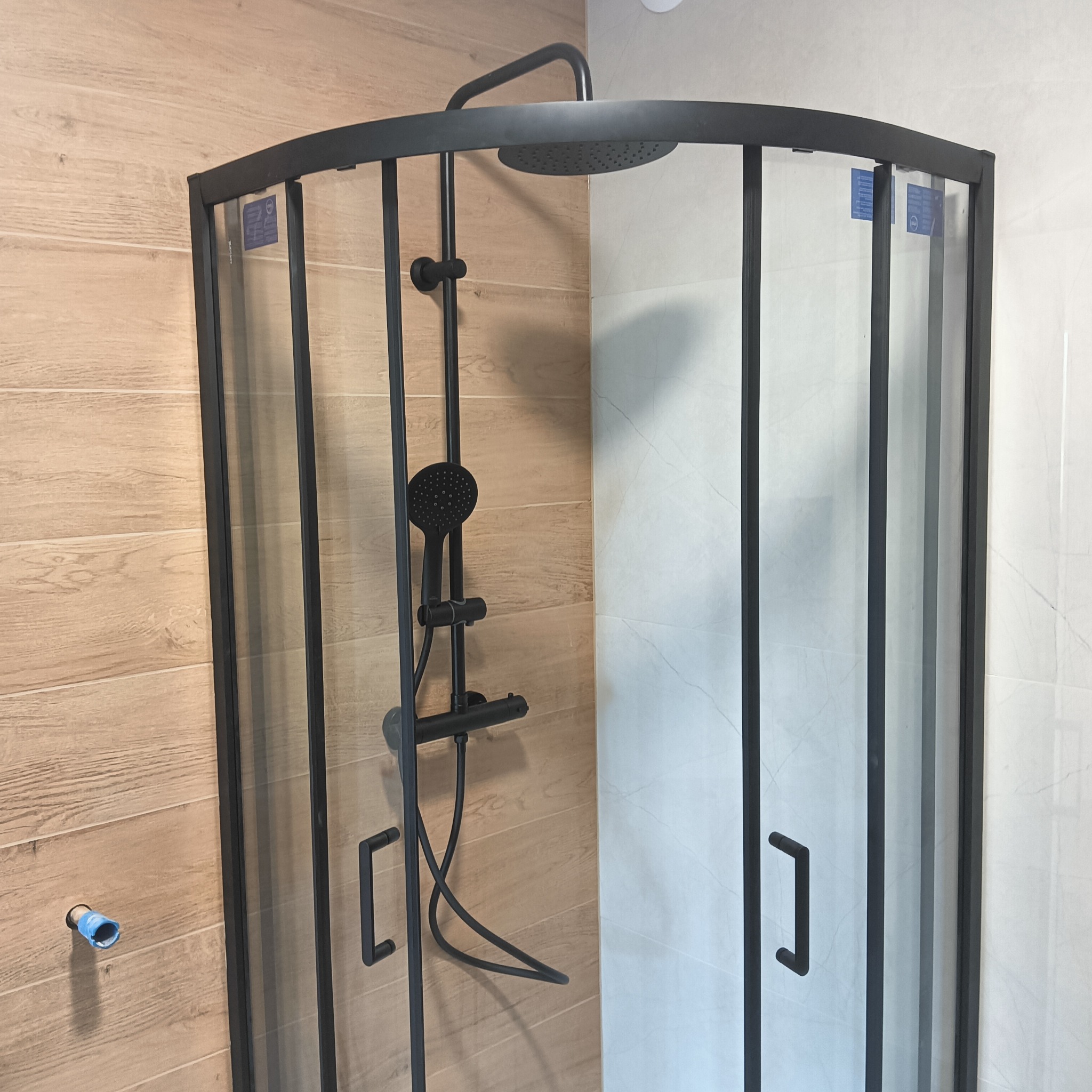 Czarna kabina prysznicowa narożna z deszczownicą na tle drewnopodobnych i marmurowych płytek w wykończonej łazience. Widoczny fragment instalacji.