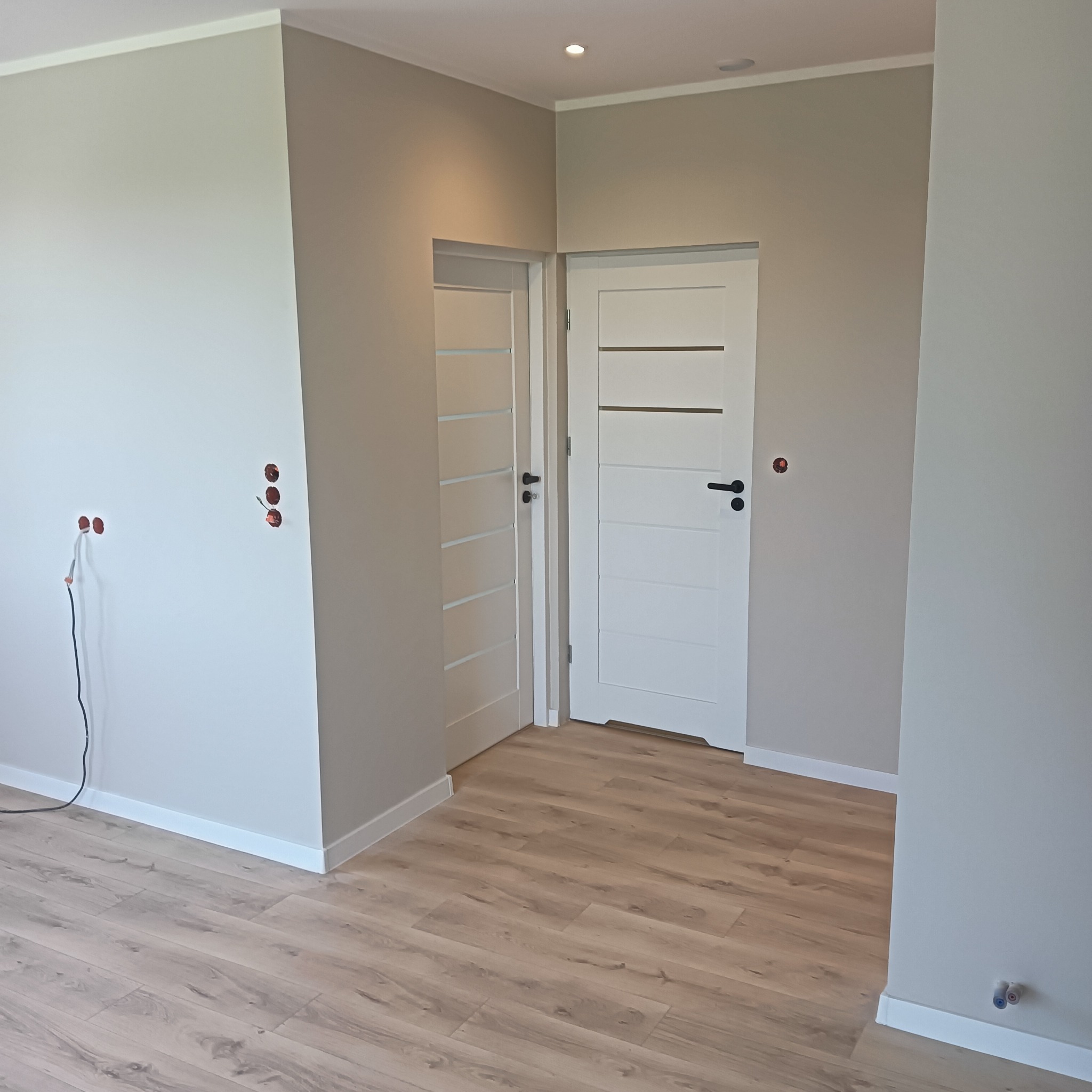 Jasne wnętrze po remoncie: dwie białe, nowoczesne drzwi z czarnymi klamkami, drewnopodobna podłoga, ściany w odcieniach szarości. Widoczne gniazdka elektryczne.