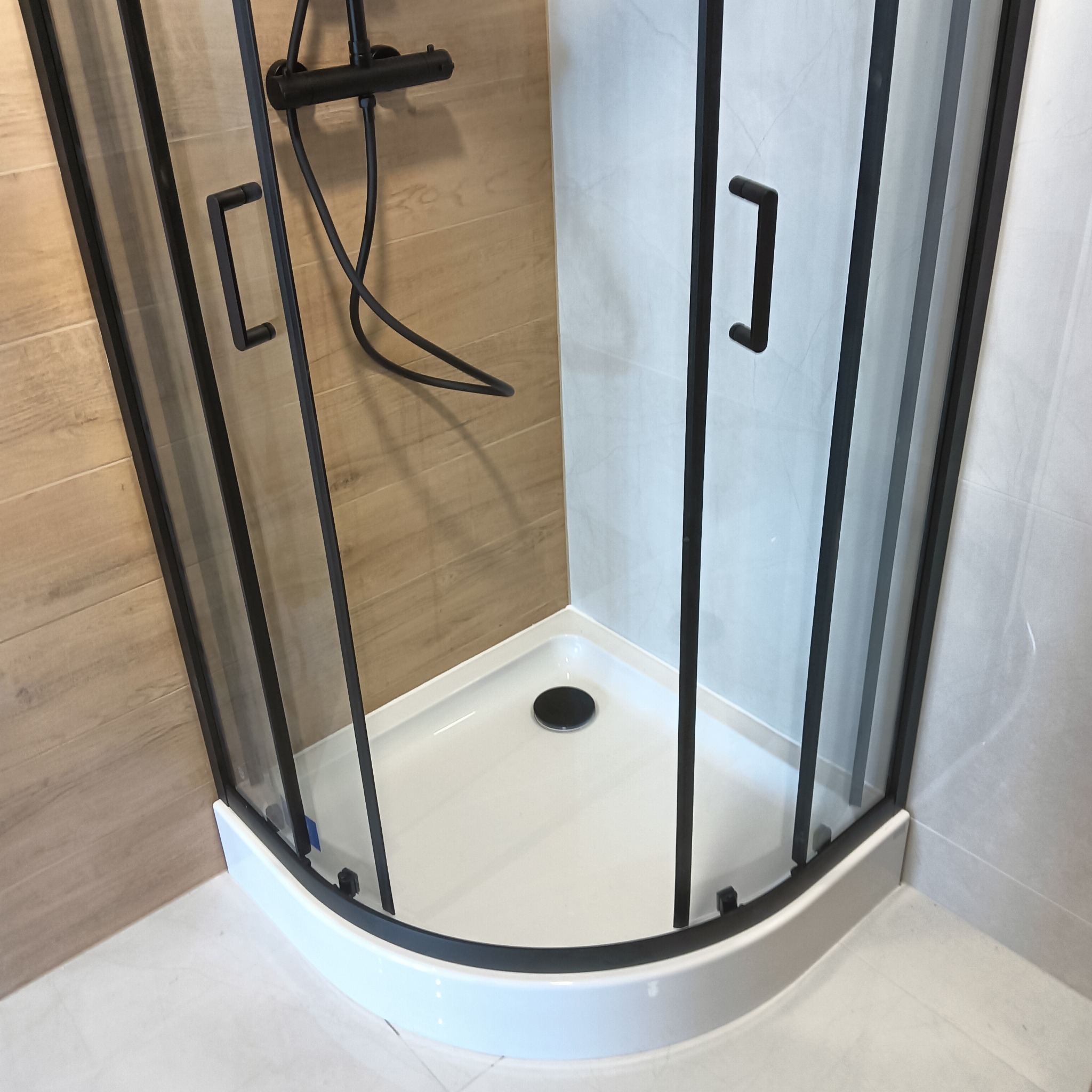 Nowoczesna kabina prysznicowa narożna z czarnymi profilami i armaturą, widok z góry, jasne płytki imitujące drewno i marmur. Minimalistyczny design.