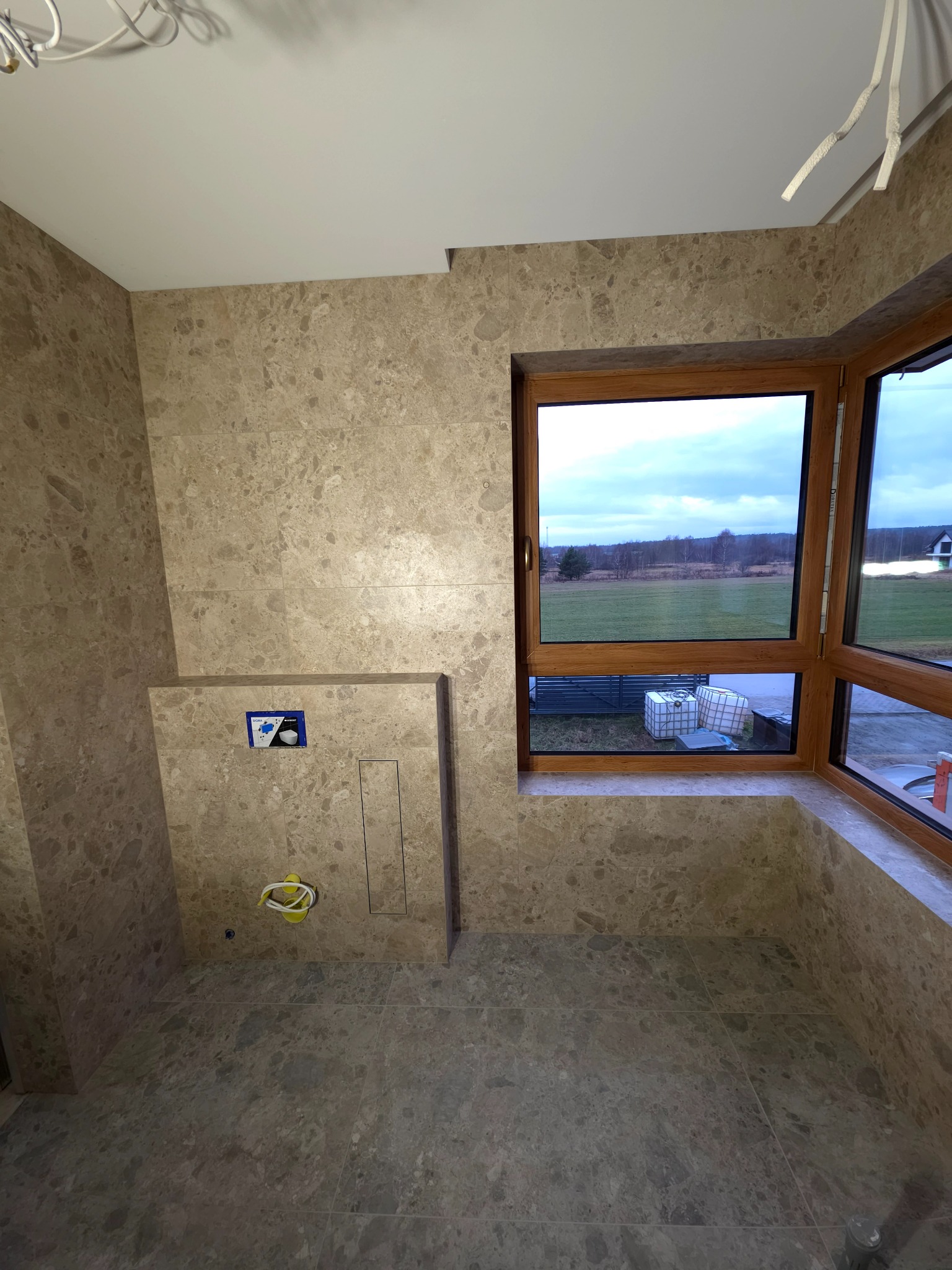 Wykończona łazienka z beżowymi płytkami imitującymi marmur, drewnianym oknem z widokiem na zewnątrz i podtynkową instalacją WC. Minimalistyczny design.