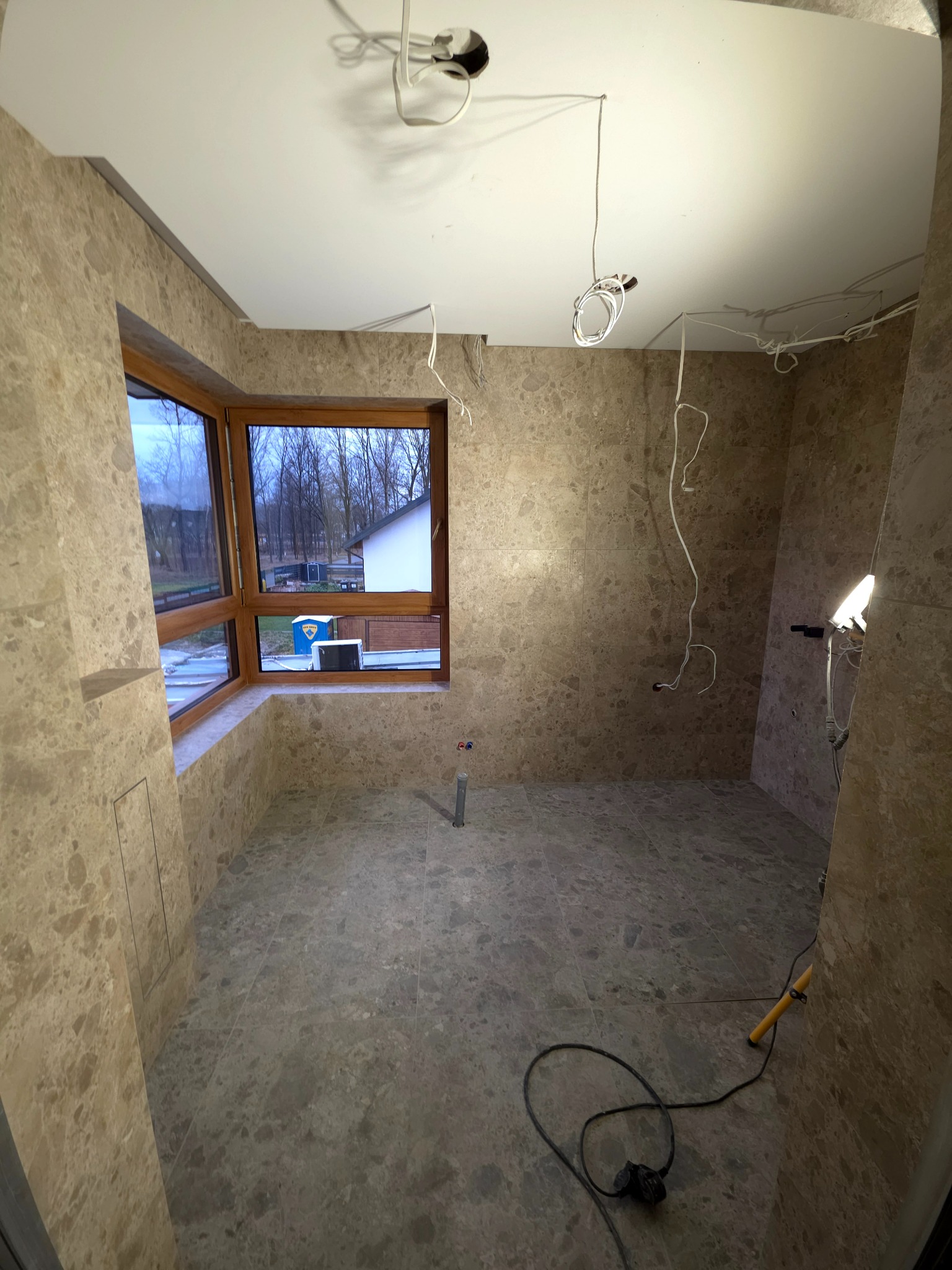 Wykończona łazienka z marmurowymi płytkami, widoczne okno z drewnianą ramą, wystające kable i narzędzia. Etap prac wykończeniowych, surowy stan.