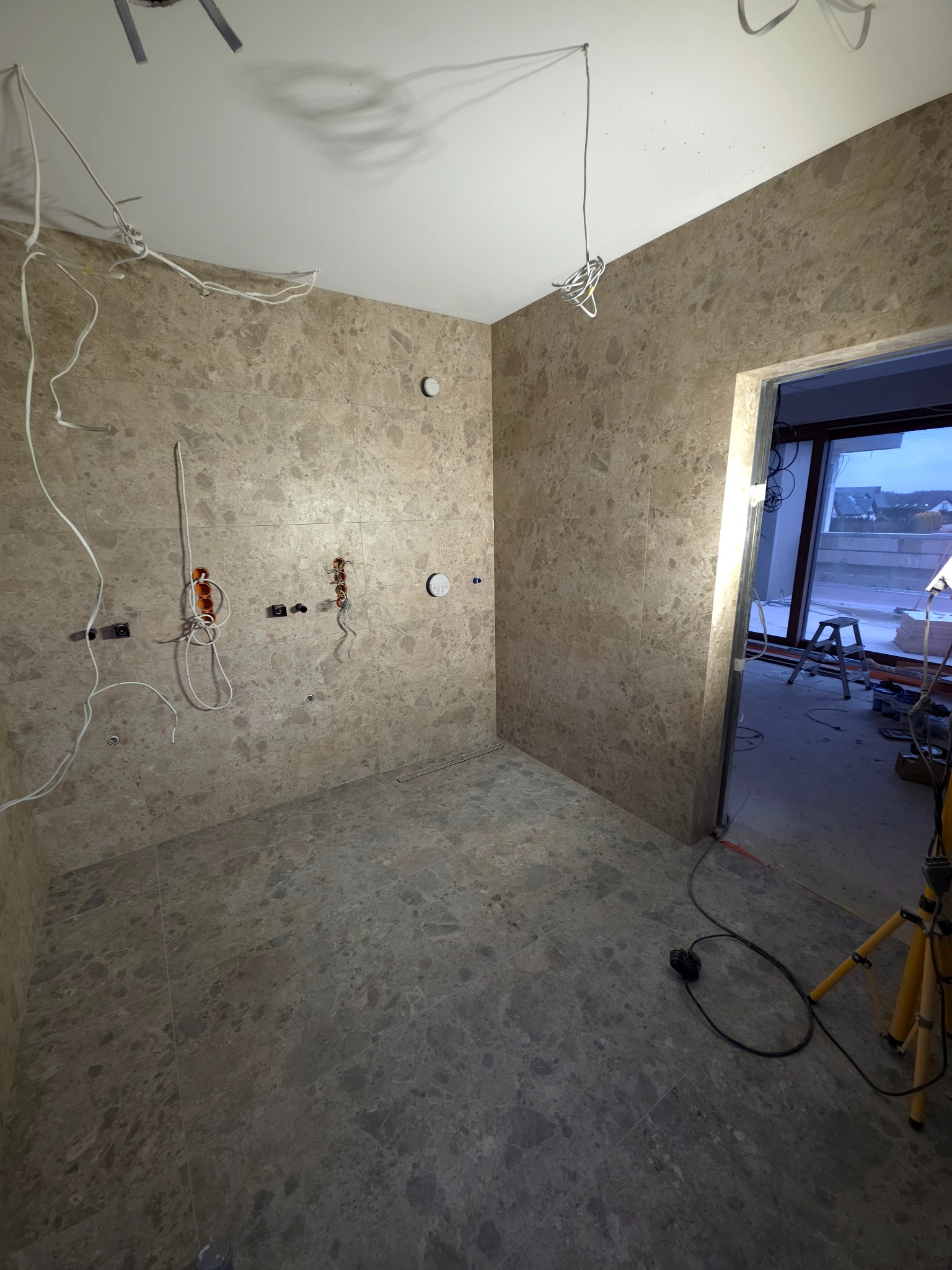 Wykończona łazienka z marmurowymi płytkami, widoczne wyprowadzone instalacje elektryczne i hydrauliczne. W tle drabina i okno z widokiem na domy.