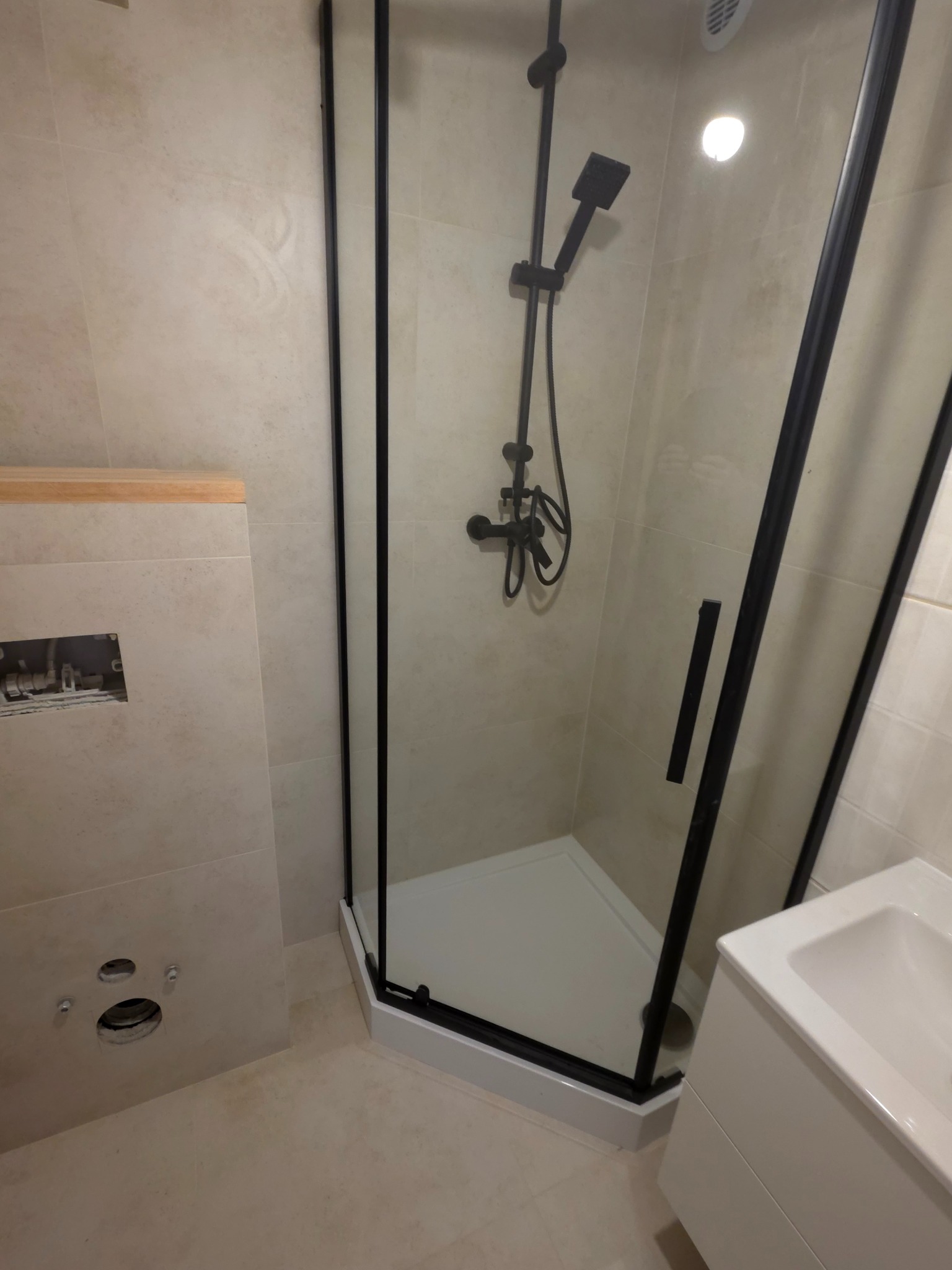 Nowoczesna kabina prysznicowa z czarnymi profilami i deszczownicą, jasne płytki, fragment umywalki. Widoczny etap prac wykończeniowych w łazience.