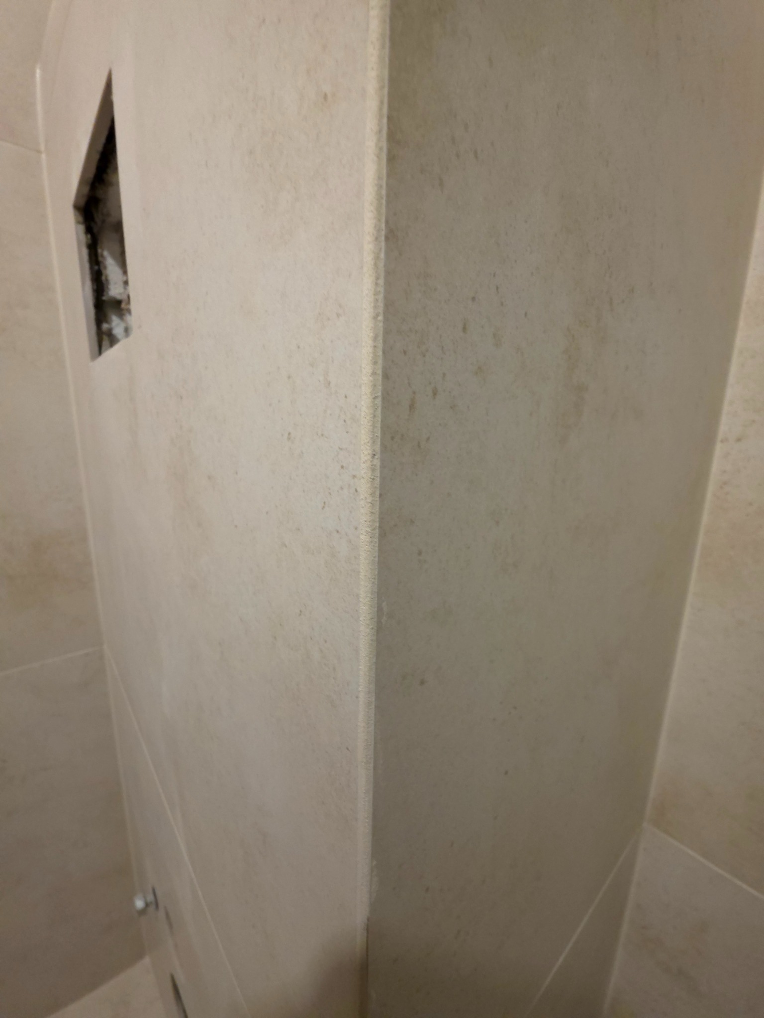 Ściana łazienki wyłożona beżowymi płytkami z widocznym otworem na instalację. Widoczny narożnik i fugi. Delikatna faktura płytek. Ujęcie z dołu.